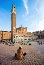Young woman sitting on the bricks of a plaza in Siena facing a tall bell tower