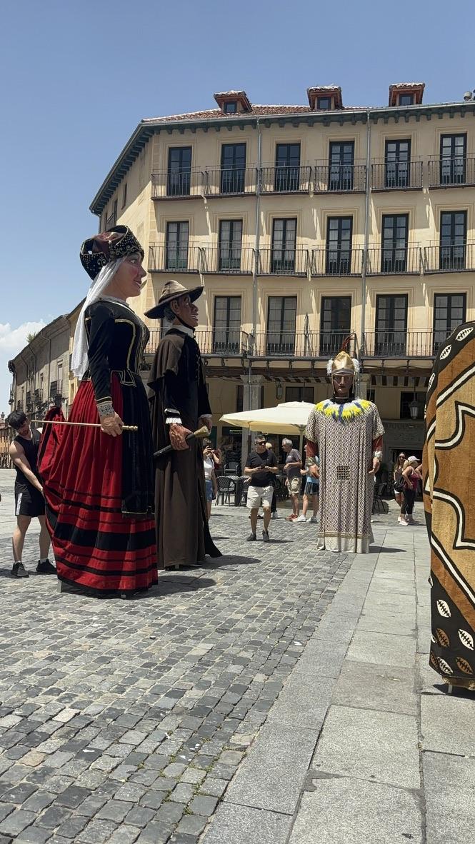 Two individuals in traditional Spanish costumes, one wearing a black and red dress with a headpiece, and the other in a brown outfit with a hat, participating in a cultural event in a sunny plaza.