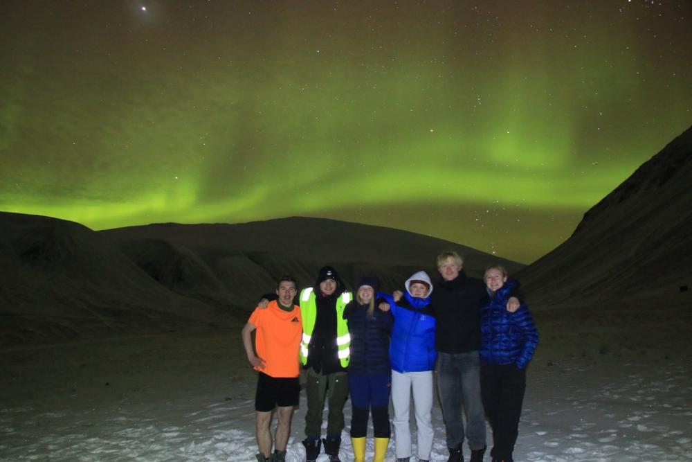 A group of six students stand together in the snow with a green tint of the northern lights in the sky behind them