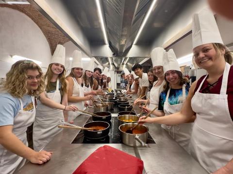 College aged students in a large kitchen wearing white aprons and tall paper white hats