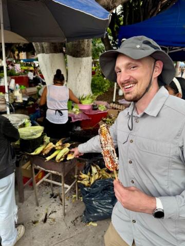 A person in a sun hat points to four cobs of corn on a griddle in the background, while holding a cob of recently-grilled corn 