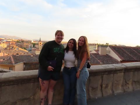 three people stand on a balcony, with city buildings in the background