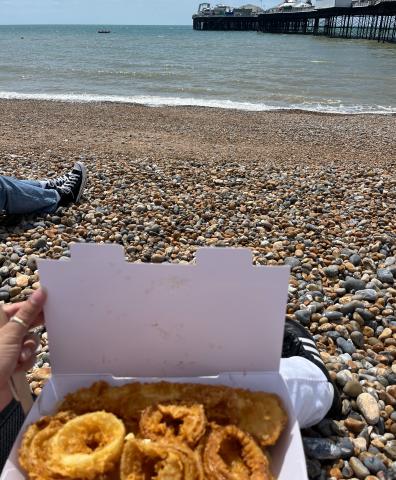 Person sitting on a pebble beach holding a box of onion rings, with a view of the ocean and Brighton Pier in the background.