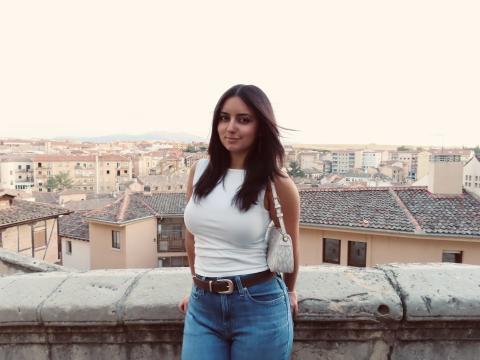 Ava stands on a balcony in blue jeans and a white shirt with a cityscape in the background
