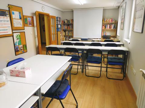 A small classroom with long white tables and blue folding chairs and a book case and projector screen in the background