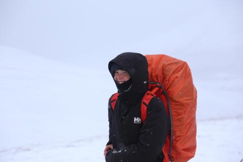 Maddy smiles at the camera with a red backpack and black coat on. there is snow in the background