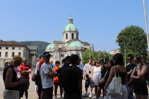 Students stand outside near a promenade of buildings, focused on one speaker