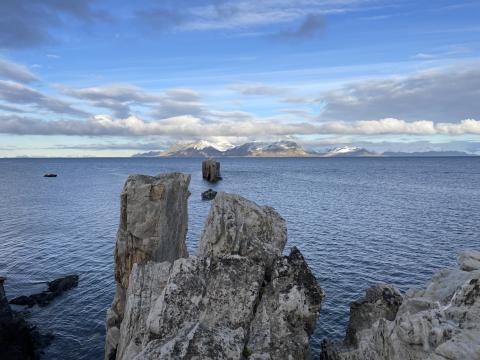 long grey rocks stick out of the ocean; there is a land formation in the distance and blue, cloudy skies