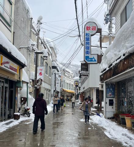 Snow-covered shopping street in Japan with people walking and storefronts on either side. A mountain is visible in the background. Signs display Japanese characters.