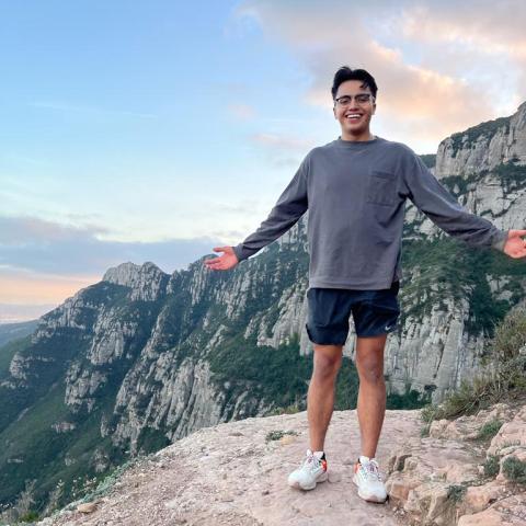 A UO student posing on the edge of a cliff against a backdrop of mountains and a sunset with his arms spread open