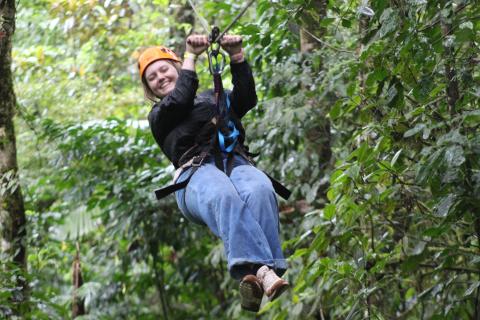 Person in a black shirt and blue jeans is ziplining, with green trees in the background