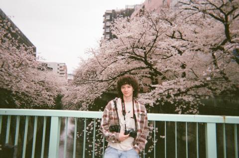 College male standing in front of a background of cherry blossom trees in Japan with a professional camera around his neck. 