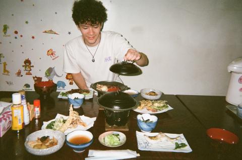 Male college student sitting at a table and opening the lid of a soup pot. There is a traditional Japanese meal laid out before him. 