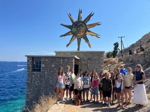 Group photo of about 15 students. They are outside by the water and have a statue of a sun behind them