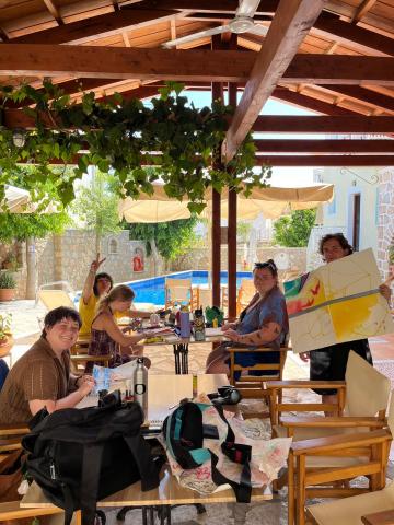 5 classmates work on painting at a long table with tall chairs; they are outside and shaded by a canopy
