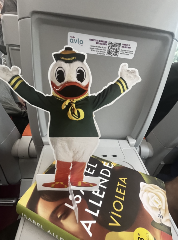 A paper cutout of the Duck with its arms up rests on an airplane tray table, a copy of a book is underneath it
