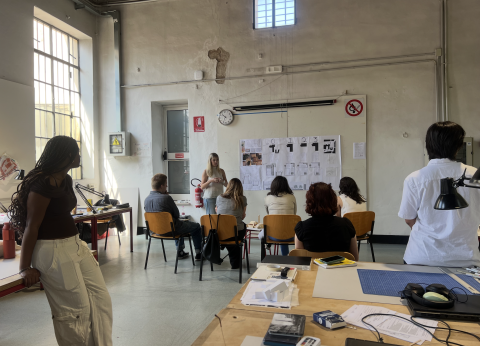 A student stands with a poster of designs with a few students sitting and watching them present