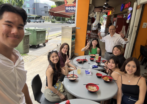 8 people sit around two small circular tables outside with plates of food in front of them