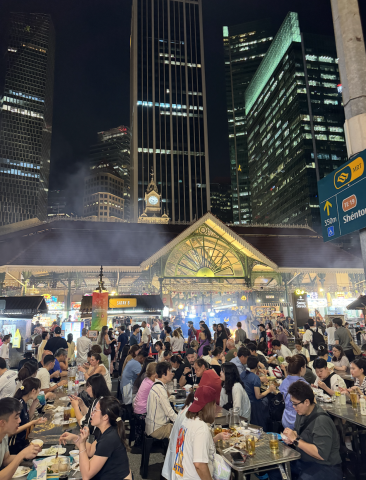 Outdoor rectangular tables are crowded with people eating, a market of food is in the background along with a nighttime city skyline