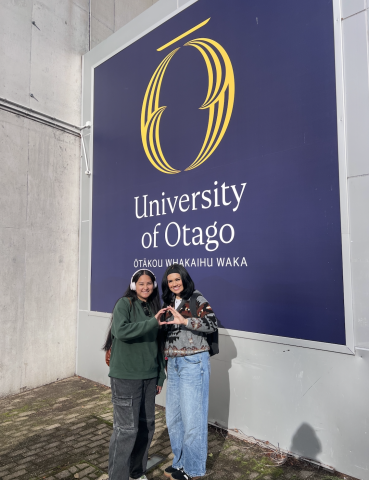 Two people pose, throwing their "O" in front of a University of Otago sign in the background