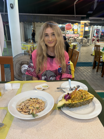 Madison is in a pink shirt and its at a table with two dishes in front of her. One has half a pineapple and the other has noodles