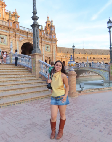 Ava stands in a yellow shirt and denim skirt with a city building and bridge behind