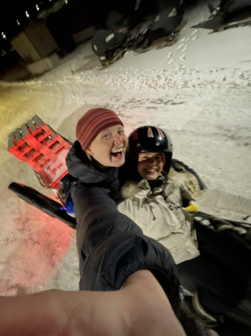 Maddy and a friend are bundled up in their coats, ear warmers, and helmets, on a sled in the snow