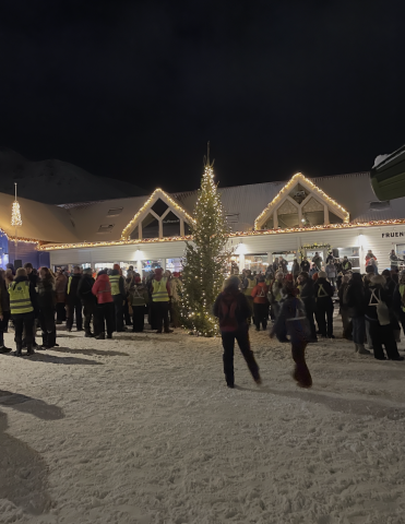 a large building is strung with lights and a lit up tree in the center of the outdoor gathering with groups of people around it