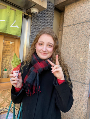 Elizabeth, dressed in a black coat and red and black scarf, poses with a peace sign in one hand and a green ice cream cone in another