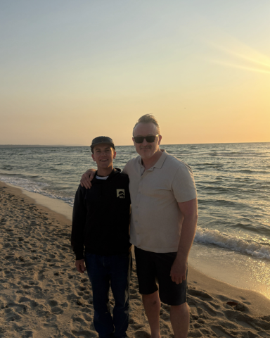 2 people stand on a beach, with the sun setting in the background