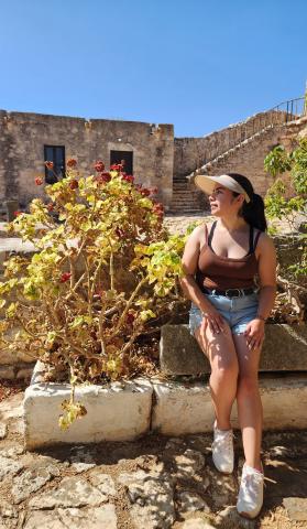 College aged woman sitting on a slab of stone next to a plant in front of an ancient stone structure in Greece