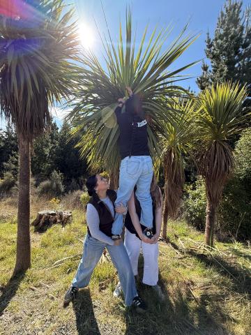 two people lift their friend up, holding them by the legs, as they reach for something up high in a tree