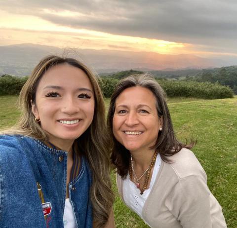 A college aged woman taking a selfie with a middle aged woman in a grass field during sunset