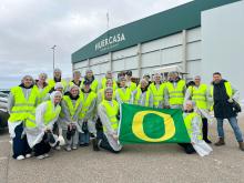 A group of twenty students wearing safety jackets and hard hats stand in front of the Huercasa factory while holding a UO flag. 