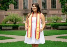 College-aged woman smiling in front of the University of Oregon's Knight Library. She is wearing a Spain flag graduation stole.