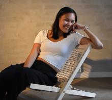Person smiling and sitting relaxed in a white folding chair, wearing a white top and black pants, in a room with a neutral toned wall.