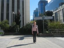 Person standing on a sunny pathway with street signs for Paseo de la Castellana and Orense in the foreground, and modern high-rise buildings in the background.