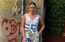 Person standing on a city street, dressed in a contemporary outfit featuring white and blue designs, accessorized with a white bag and sneakers. Graffiti is visible on the wall beside them.