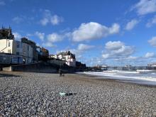 View of a pebble beach with a few individuals in the distance, facing traditional seafront buildings under a blue sky with scattered clouds.