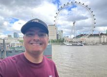 A person smiling in front of the London Eye and the River Thames on a partly cloudy day.