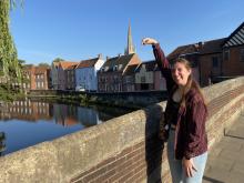 Person pretending to touch the spire of Norwich Cathedral from across the river, with colorful houses lining the opposite bank.