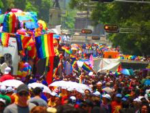 Colorful Pride parade in progress with a crowd of people and various rainbow flags and decorations in a sunny urban setting.
