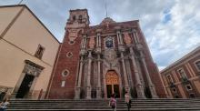 Exterior view of the Templo de San Francisco, an ornate colonial-style church with intricate façade details, under a cloudy sky. People are seen walking on the steps in front.