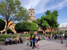 People relaxing and walking in a vibrant public square with lush green trees and a large manicured bush, with a view of a historic church tower under a clear blue sky.