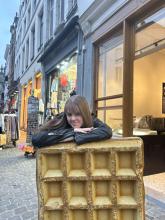 A person resting their elbows on a large golden waffle statue on a street, with shops in the background.