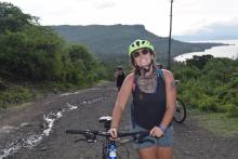 Person smiling at the camera while standing with a bicycle on a dirt path in a natural setting, wearing a helmet and sunglasses. There's a scenic view of hills and a lake in the background.