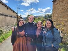 Four friends smiling and posing together on a sunny day with a scenic background of a country road and stone walls.