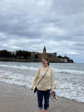 Person standing on a sandy beach with waves at their feet, smiling with the old town of Gijón, Spain, featuring the St. Peter's Church in the background under a cloudy sky.