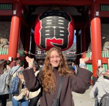 A person smiling and posing in front of the Kaminarimon gate at Senso-ji Temple, Tokyo, with a large red lantern overhead.