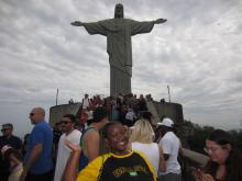 Person wearing a Brazil T-shirt smiling in front of the Christ the Redeemer statue in Rio de Janeiro, with other visitors around.